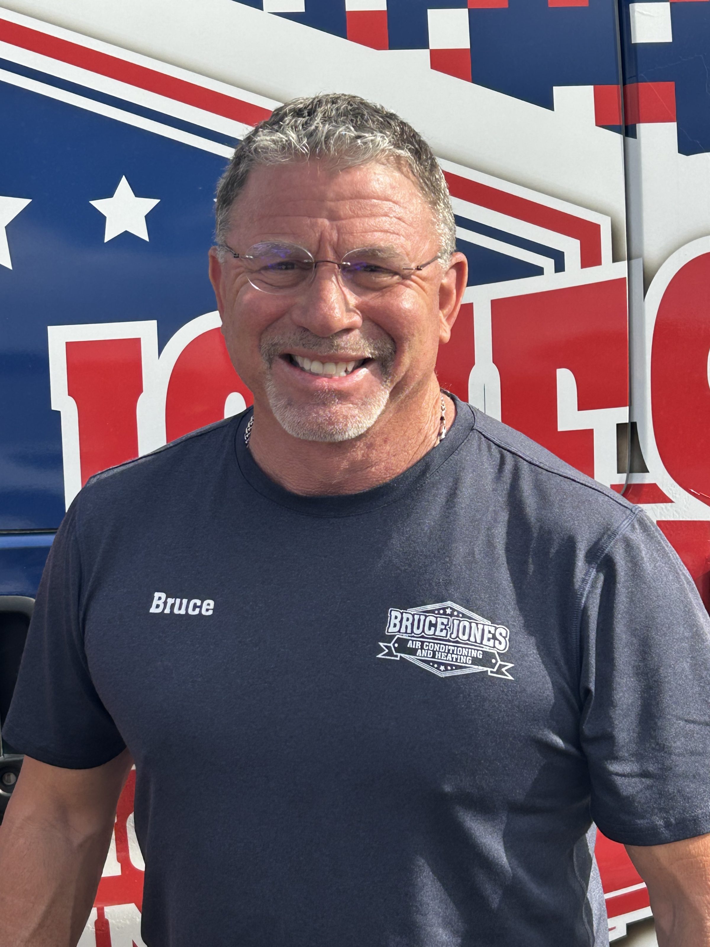 Smiling man wearing glasses and a dark HVAC company shirt in front of red, white, and blue truck graphics
