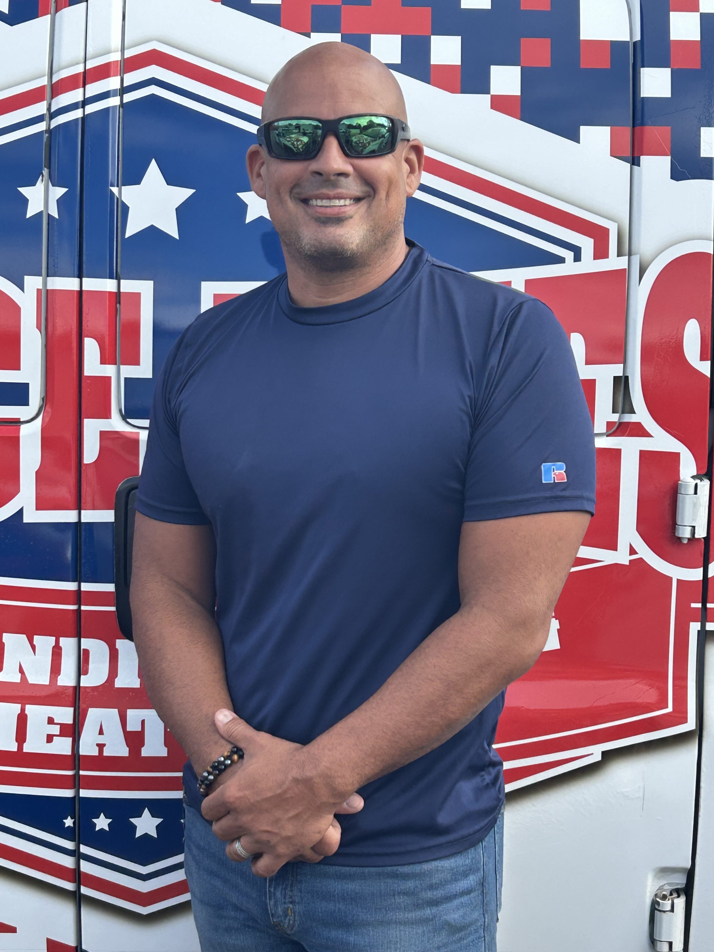 A smiling man in sunglasses and a navy t-shirt stands in front of a red, white, and blue patriotic vehicle decal.