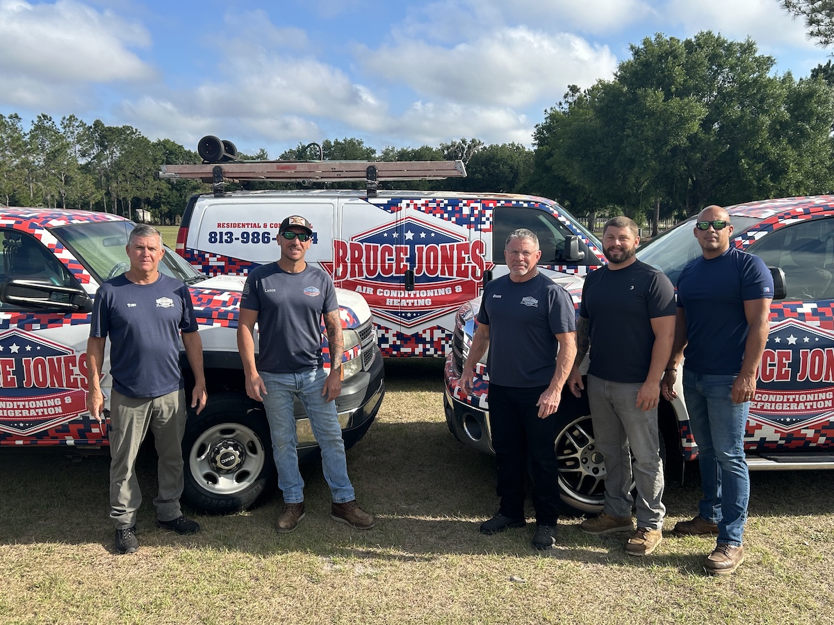 Five HVAC technicians standing in front of Bruce Jones Air Conditioning & Heating service vans with patriotic wrap.