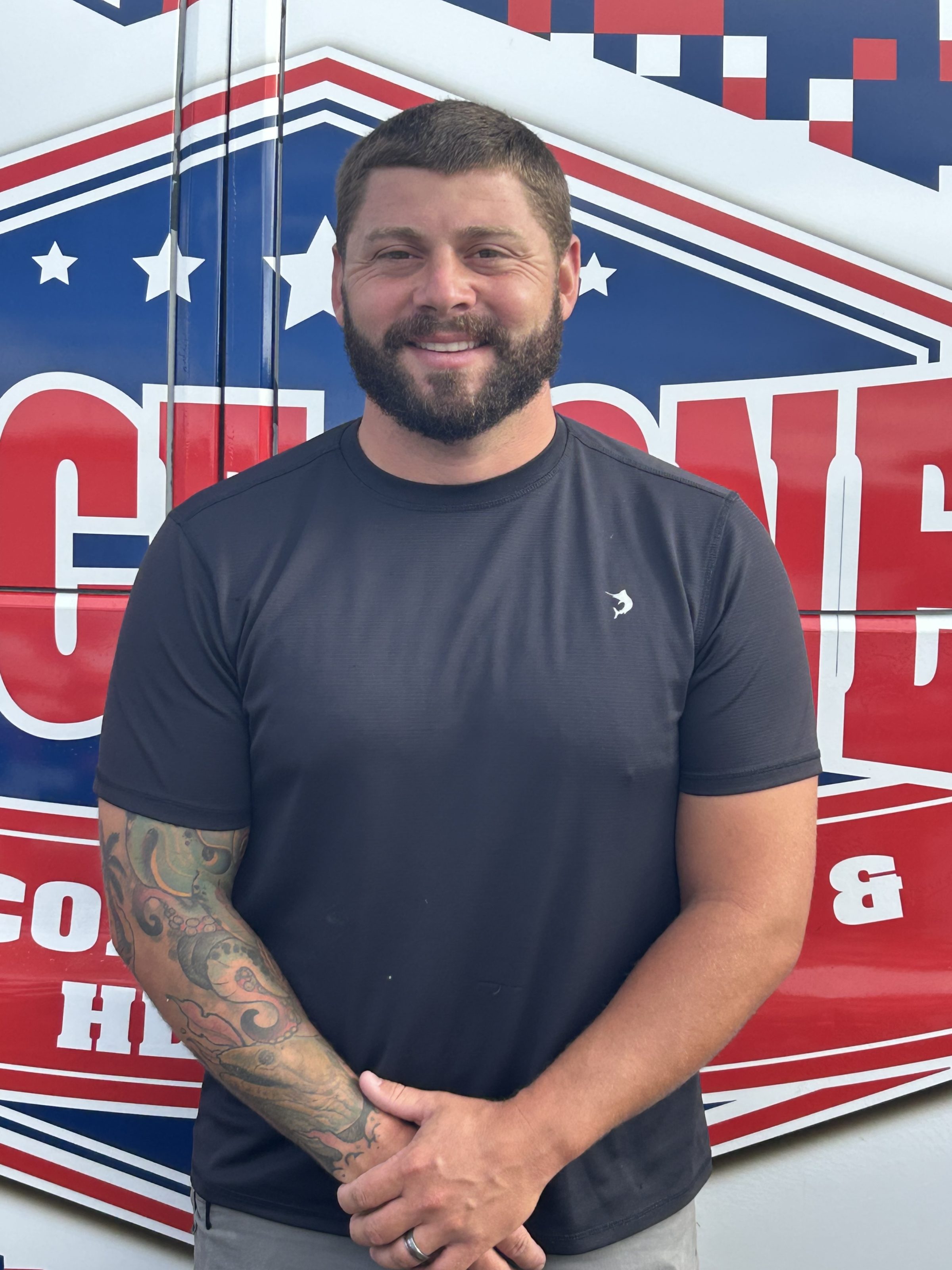Smiling bearded man in black shirt with tattooed arm standing in front of red, white and blue patriotic graphic