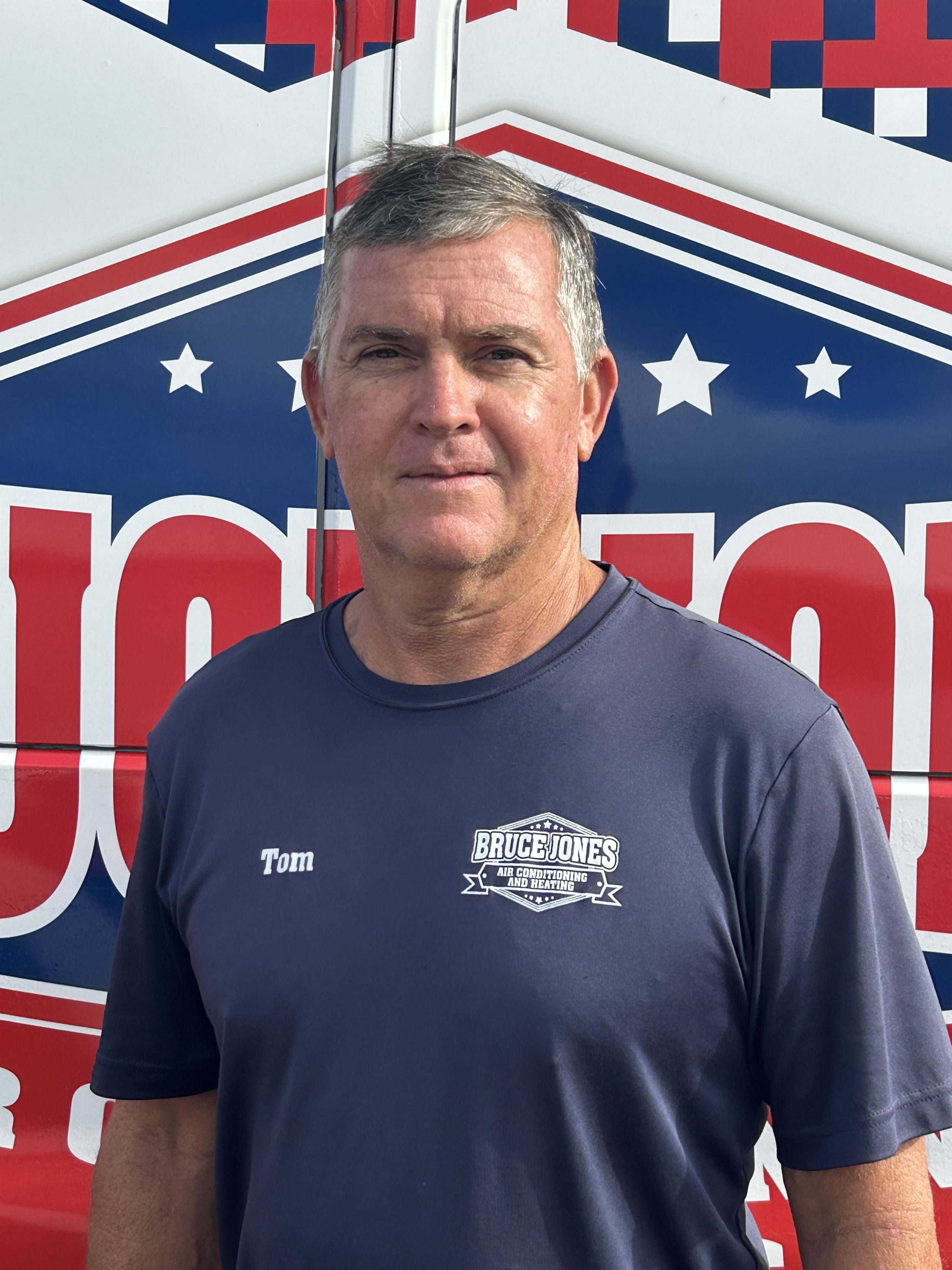 A man wearing a Bruce Jones Air Conditioning & Heating shirt standing in front of a red, white and blue company van